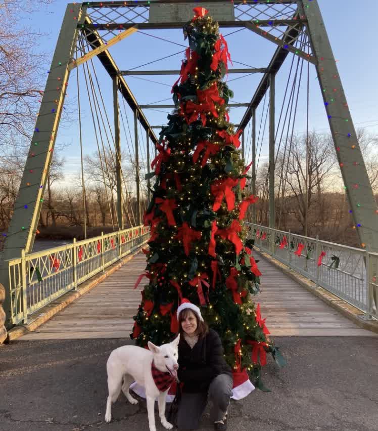 Margaret in a Santa hat kneeling beside Ella, who is wearing a red plaid bandana, in front of a tall decorated Christmas tree on a bridge
